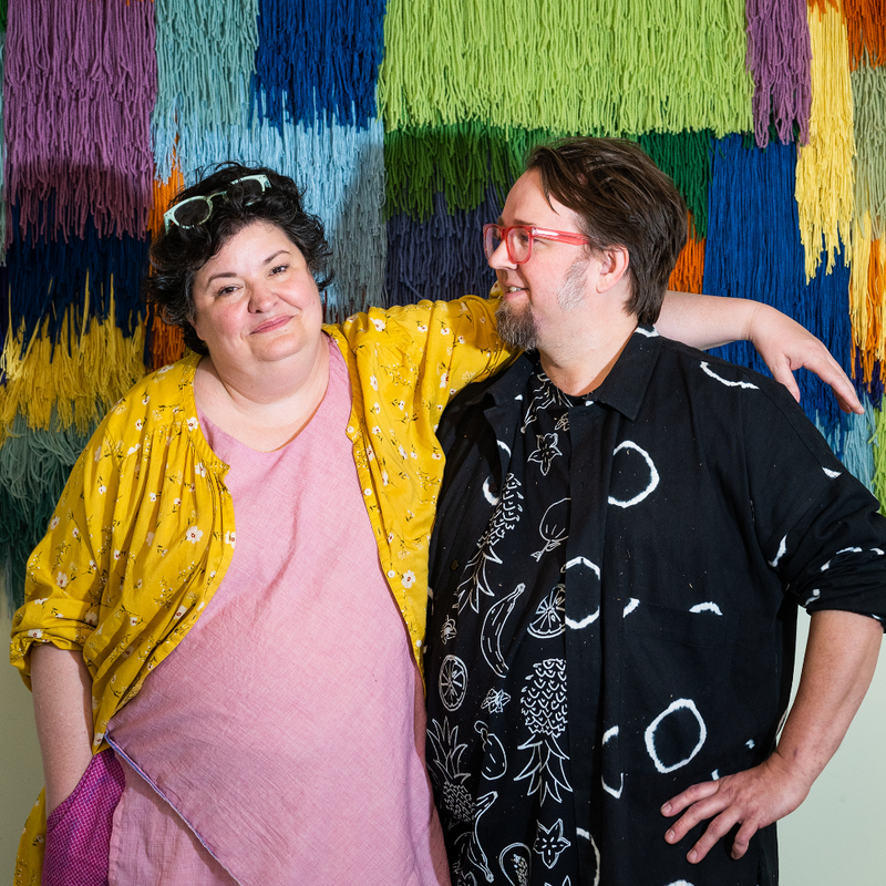 Tim Eads and Tiernan Alexander standing in front of a colorful textured wall with a pink lamp above.