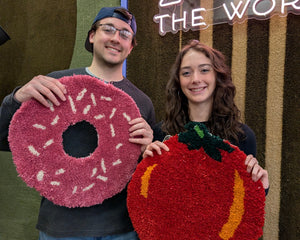 A man and a woman stand together holding large, plush, shaggy fruit-shaped wall art pieces. The man holds a pink donut with white sprinkles, and the woman holds a red tomato with a green stem and yellow accents.