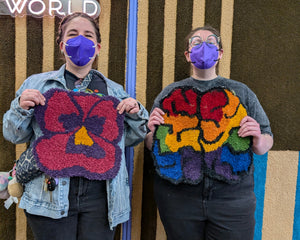 Two people wearing purple face masks stand in front of a decorative wall, each holding a colorful, hand-knotted or tufted textile square. The person on the left holds a purple and red floral design with a yellow center, and the person on the right holds a multicolor abstract design.