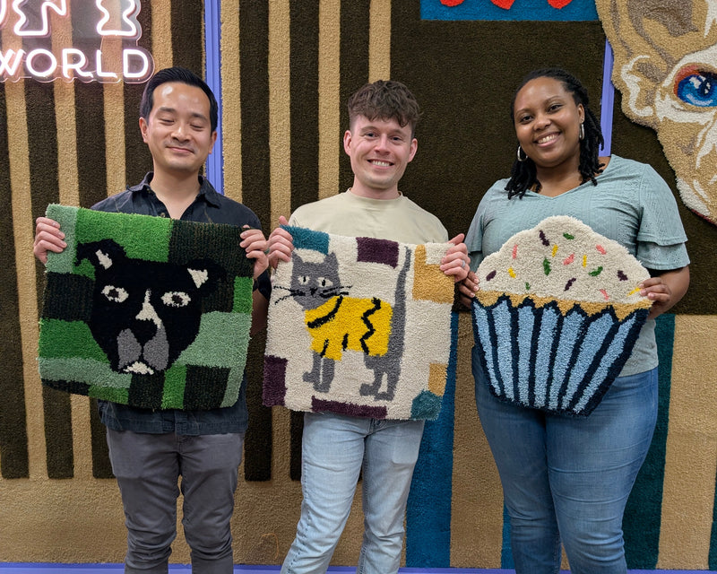 Two people wearing purple face masks stand in front of a decorative wall, each holding a colorful, hand-knotted or tufted textile square. The person on the left holds a purple and red floral design with a yellow center, and the person on the right holds a multicolor abstract design.
