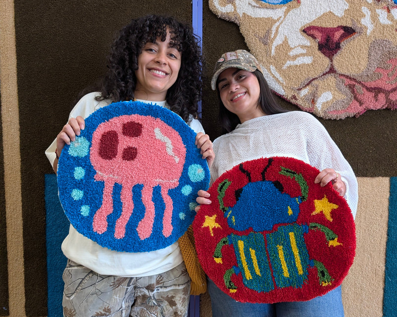 Two women holding up handmade wool rugs in front of a colorful animal-themed wall. The rug on the left is dark blue and black with pink and gray cat motifs, and the rug on the right is black and beige with a simple cat face design.