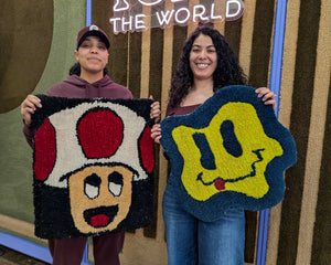 Two women standing in front of a 'TUF THE WORLD' sign, holding up hand-pile-rupee character rugs. The woman on the left holds a Toad-themed rug, and the woman on the right holds a yellow smiley face rug.
