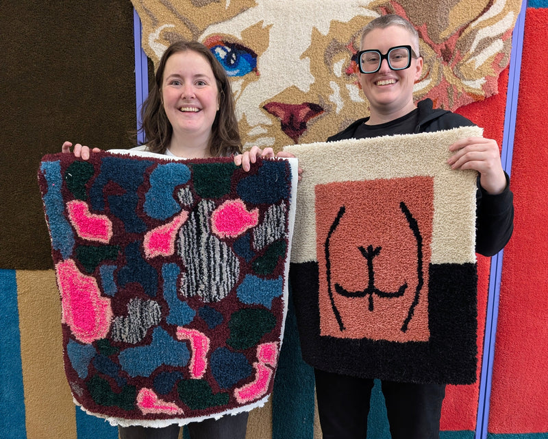 Two women holding up handmade wool rugs in front of a colorful animal-themed wall. The rug on the left is dark blue and black with pink and gray cat motifs, and the rug on the right is black and beige with a simple cat face design.